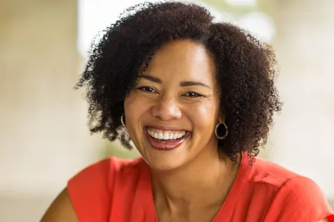 Cheerful woman with curly hair smiling warmly, wearing a vibrant orange blouse in a natural outdoor setting.