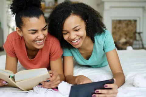 Two smiling teenage girls lying on bed reading a book and using a tablet together in a cozy room.