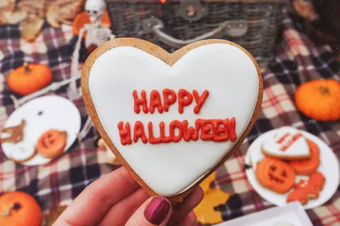 Hand holding heart-shaped cookie with Happy Halloween icing, autumn picnic setup with pumpkins and Halloween treats.