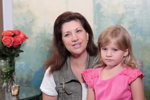 Mother and daughter sitting indoors with vase of orange roses on glass table beside them.