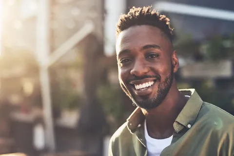 Smiling man outdoors in natural light, wearing a casual shirt, showcasing positive attitude and warmth.