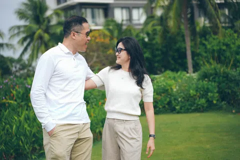 Couple in casual beige and white outfits smiling and walking in a green garden with tropical plants.