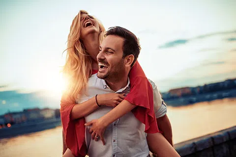 Happy couple laughing and enjoying a piggyback ride by the waterfront at sunset with cityscape in the background