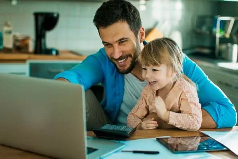 Father and daughter smiling while using laptop and tablet together at a wooden kitchen table.