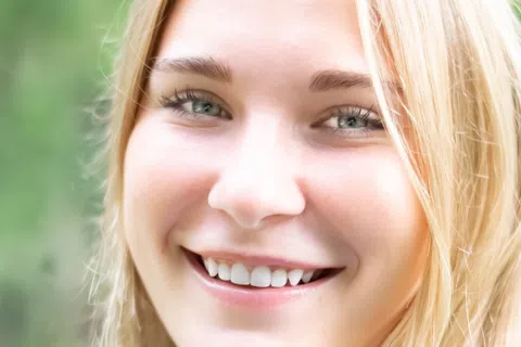 Close-up of a smiling young woman with blonde hair and blue eyes against a blurred green background