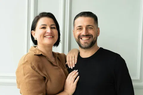 Smiling couple standing close with woman in brown shirt and man in black sweater against white wall.