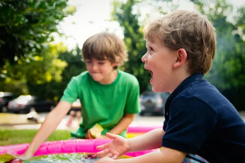Two young boys playing happily in a pink inflatable pool outdoors on a sunny day.