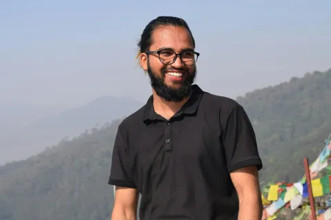 Smiling bearded man wearing glasses and black polo shirt outdoors with mountains and prayer flags in background