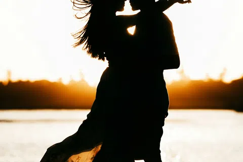 Silhouette of a couple embracing by the water during a golden sunset with flowing dress detail.