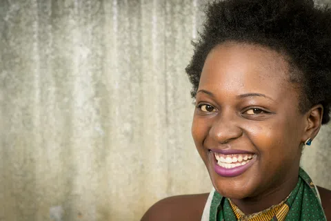 Smiling woman with short curly hair wearing purple lipstick and a green patterned top against a textured background