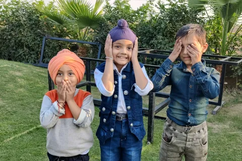 Three children outdoors playfully covering ears, eyes, and mouth in a garden with greenery and palm trees.