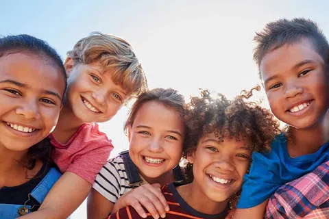 Five happy children smiling together outdoors against a bright blue sky.