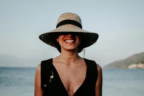 Smiling woman wearing a wide-brimmed hat by the beach during sunset.