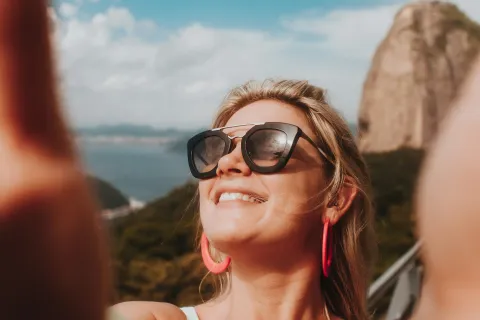 Smiling woman wearing sunglasses and pink earrings taking a selfie outdoors with a mountain and blue sky background