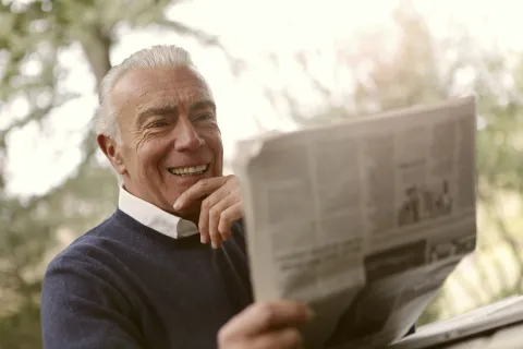 Smiling elderly man in navy sweater reading a newspaper outdoors with blurred greenery background.