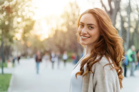 Smiling woman with long reddish hair walking in a sunlit park, surrounded by blurred people in the background.