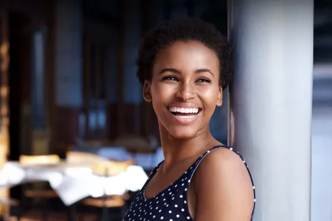 Smiling woman in polka dot dress, leaning against a pole in a cozy outdoor setting.