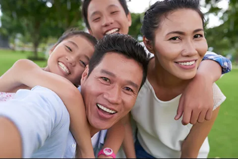 Happy Asian family of four smiling and taking a selfie outdoors in a park with green grass and trees.