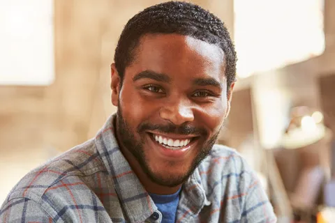 Smiling young man in a casual checkered shirt, seated in a bright modern workspace.