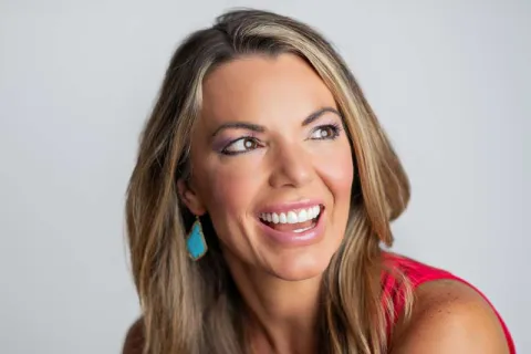 Smiling woman with long blonde hair wearing a red top and turquoise earrings looking to the side.