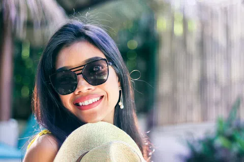 Smiling woman wearing sunglasses and holding a woven hat in a sunlit outdoor setting with blurred background.