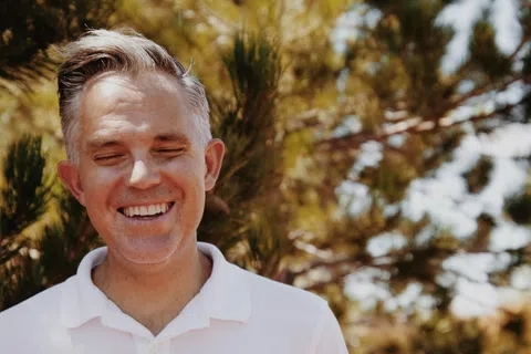 Smiling middle-aged man with gray hair wearing a white polo shirt outdoors with trees in background