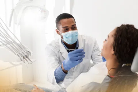 Dentist wearing mask and gloves examining a female patient's teeth in a bright dental clinic.