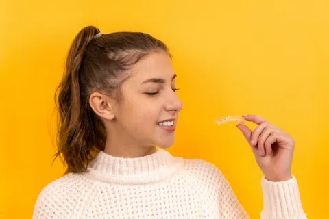 Young woman in a white sweater holding and smiling at a clear dental aligner against a yellow background.