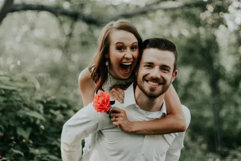 Happy couple outdoors, woman playfully hugging man from behind holding a red flower in green natural setting