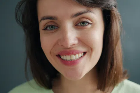 Close-up portrait of a smiling woman with green eyes and brown hair against a gray background.