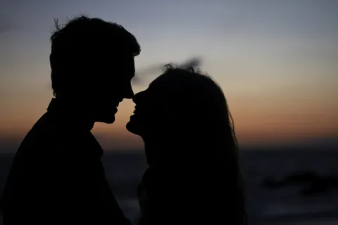 Silhouetted couple leaning in for a kiss at sunset by the ocean shore with a colorful sky.