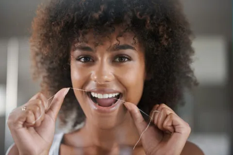 Smiling woman with curly hair using dental floss for oral hygiene in a bright bathroom setting.