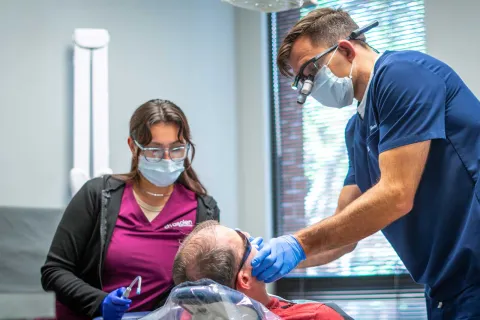Dentist and assistant wearing masks and gloves treating a patient in a dental clinic with modern equipment.