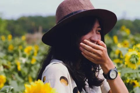 Woman laughing amidst vibrant sunflowers wearing a hat and a patterned shirt.