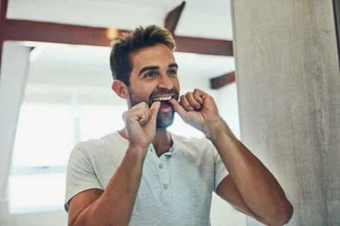 Man smiling while flossing his teeth in a bright bathroom mirror.
