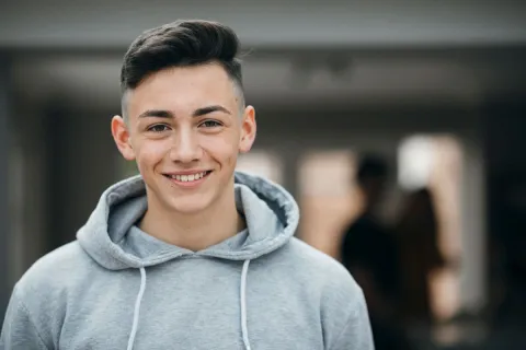 Smiling young man in a gray hoodie, indoors with a blurred background of people.