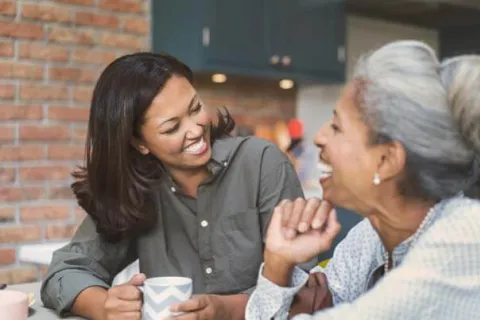 Two women laughing and enjoying coffee together in a cozy kitchen setting.