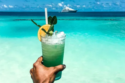 Hand holding a green cocktail with lemon garnish against turquoise ocean and yacht on horizon under blue sky.