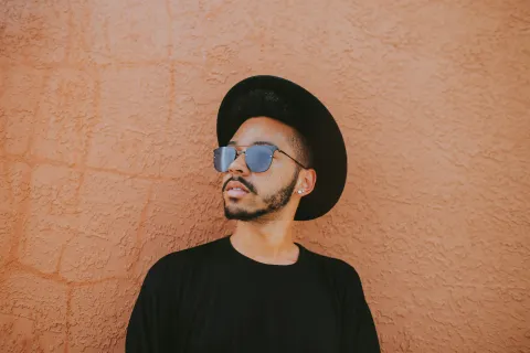 Stylish man wearing sunglasses and a black hat posing against textured orange wall, looking away thoughtfully.