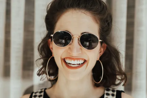Smiling woman in round sunglasses and checkered overalls, posing against draped backdrop.
