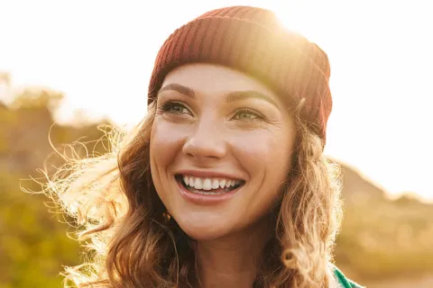 A smiling woman wearing a beanie and a green and white jacket, with sunlight glowing behind her.