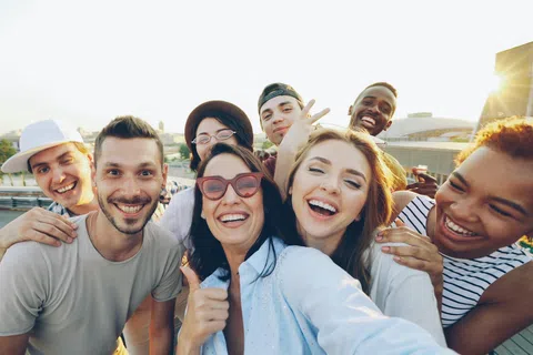 Group of diverse young adults smiling and taking a selfie outdoors on a sunny day at sunset.