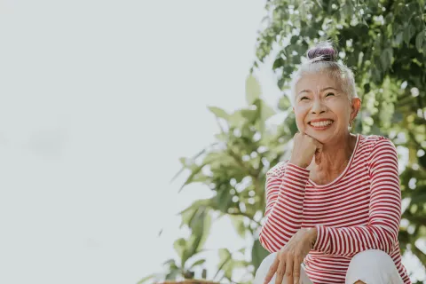 Smiling senior woman sitting outdoors in a striped shirt with greenery in the background.