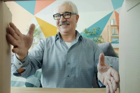 Smiling elderly man unboxing package with hands open in geometrically decorated room with plants and shelves.