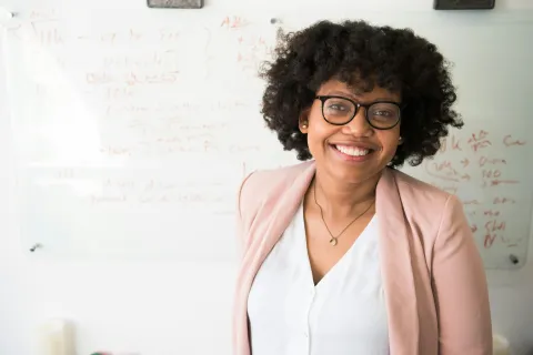 Smiling woman with glasses and curly hair standing in front of a whiteboard with notes and diagrams.