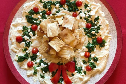 Festive holiday hummus platter decorated with cherry tomatoes, parsley, pita chips, and red pepper bow on a plate.