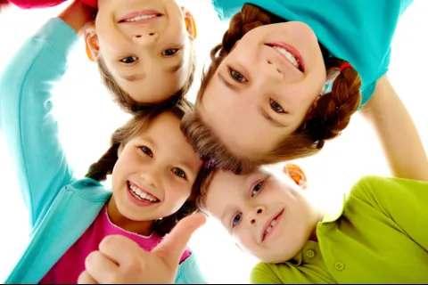 Four smiling children in colorful clothes huddled in a circle giving thumbs up and looking down at the camera.