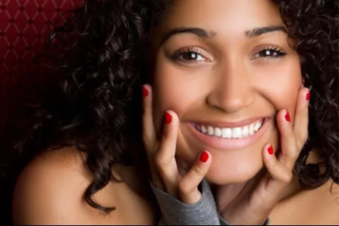 Close-up of a smiling woman with curly hair and red nail polish gently touching her face.