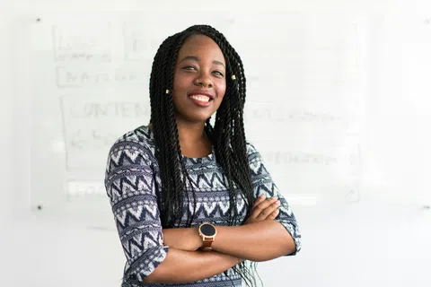 Confident young woman with braided hair standing with arms crossed in front of a whiteboard in an office.