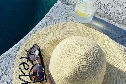 Straw sunhat with sunglasses by a poolside next to a cold drink with lemon and straw on hot sunny day.